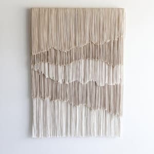 May include: A rectangular wall hanging composed of numerous vertical cotton cords arranged in four distinct horizontal layers. The cords transition in color from a light beige at the top to a gradient of tan and cream shades toward the bottom.