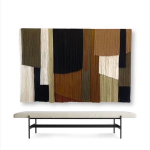 May include: A beige upholstered bench with a black metal frame sits in front of a large abstract wall hanging. The wall hanging is made of woven strips of fabric in shades of brown, green, white, and black.