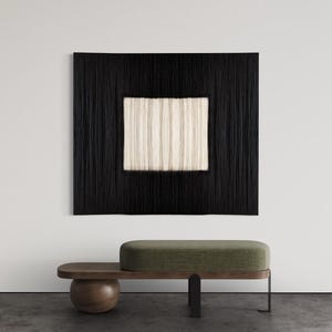 Könnte beinhalten: Ein schwarzer gewebter Wandbehang mit einem zentralen Quadrat aus vertikalen cremefarbenen Fasern. Darunter eine Bank mit grün gepolsterter Sitzfläche und einem Holzsockel mit abgerundetem Ende und einem schwarzen Metallbein.