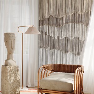 Op de afbeelding: Een lichtbruine rieten fauteuil met een beige kussen staat in een kamer met witte gordijnen en een macram&eacute; wandkleed. Een stenen sculptuur van een hoofd staat op een stenen sokkel.