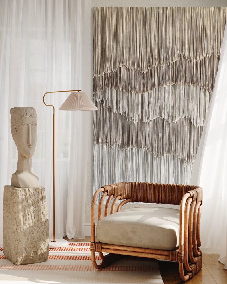 Op de afbeelding: Een lichtbruine rieten fauteuil met een beige kussen staat in een kamer met witte gordijnen en een macram&eacute; wandkleed. Een stenen sculptuur van een hoofd staat op een stenen sokkel.