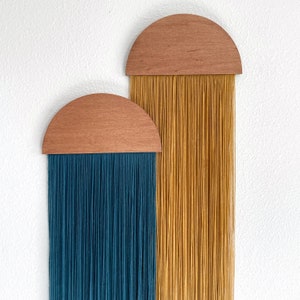 Könnte beinhalten: Zwei halbkreisförmige Wandbehänge aus Holz mit langen Garnsträngen, die herunterhängen. Der obere Halbkreis hat gelbes Garn und der untere Halbkreis hat blaugrünes Garn.