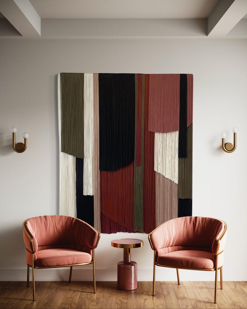 May include: Two pink velvet armchairs with gold metal frames sit facing each other in front of a large abstract wall hanging made of colorful yarn. A small pink side table sits between the chairs.