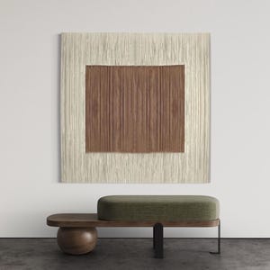 Könnte beinhalten: Moderne Innenszene mit einer strukturierten Wandbehänge mit hellbeigem Rand und braunem Zentrum. Darunter eine Bank mit Holzsockel, grün gepolsterter Sitzfläche und dunklem Metallbein.