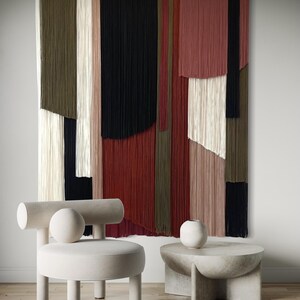 May include: A white round chair with a cylindrical back and a round cushion. The chair is in front of a wall hanging made of colorful yarn strands. A concrete side table with a round top and a white ceramic vase sits next to the chair.
