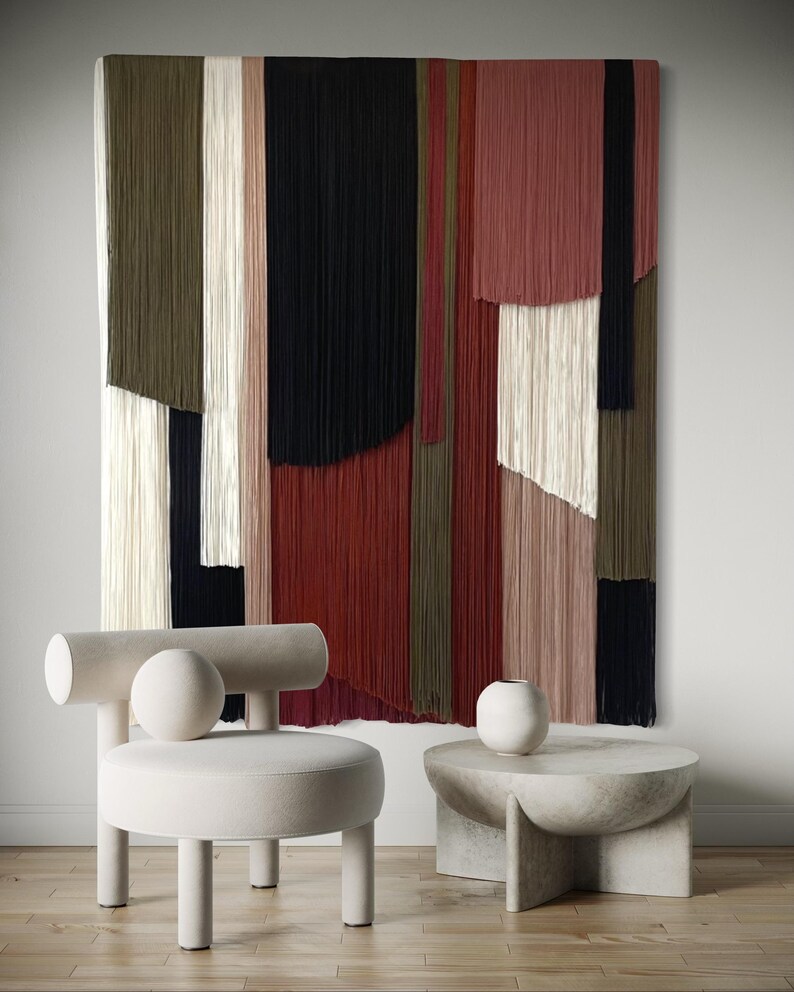 May include: A white round chair with a cylindrical back and a round cushion. The chair is in front of a wall hanging made of colorful yarn strands. A concrete side table with a round top and a white ceramic vase sits next to the chair.