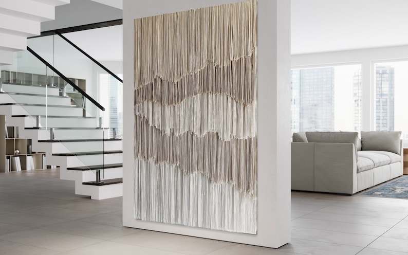 Op de afbeelding: Een groot wandkleed gemaakt van geweven garen in beige en witte tinten. Het garen is gerangschikt in verticale lijnen, waardoor een textuurrijk en abstract ontwerp ontstaat.
