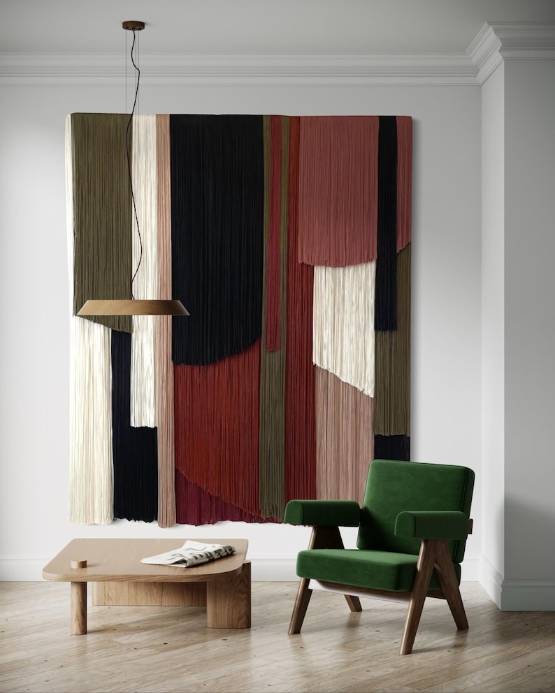 May include: A green velvet armchair with wooden legs sits in front of a large wall hanging made of colorful yarn strands. A wooden coffee table with a magazine on top sits in front of the chair.