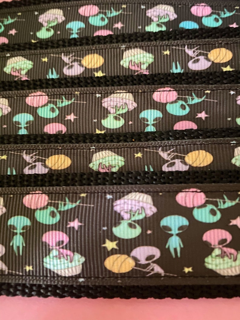K&ouml;nnte beinhalten: Schwarzes Band mit einem sich wiederholenden Muster aus pastellfarbenen Au&szlig;erirdischen, UFOs, Sternen und Planeten. Das Band hat ein strukturiertes, geripptes Aussehen mit einem schwarzen Rand. Die Au&szlig;erirdischen sind in Gr&uuml;n-, Lila- und Blaut&ouml;nen gehalten.