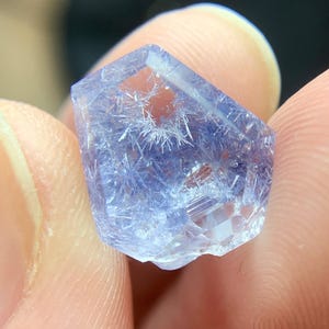 以下が含まれることがあります： 内部に針状のインクルージョンを持つ、ファセット加工された半透明の青い結晶。 結晶は幾何学的な形状をしており、2本の指の間に挟まれています。 色は、紫がかった淡い氷のような青色です。