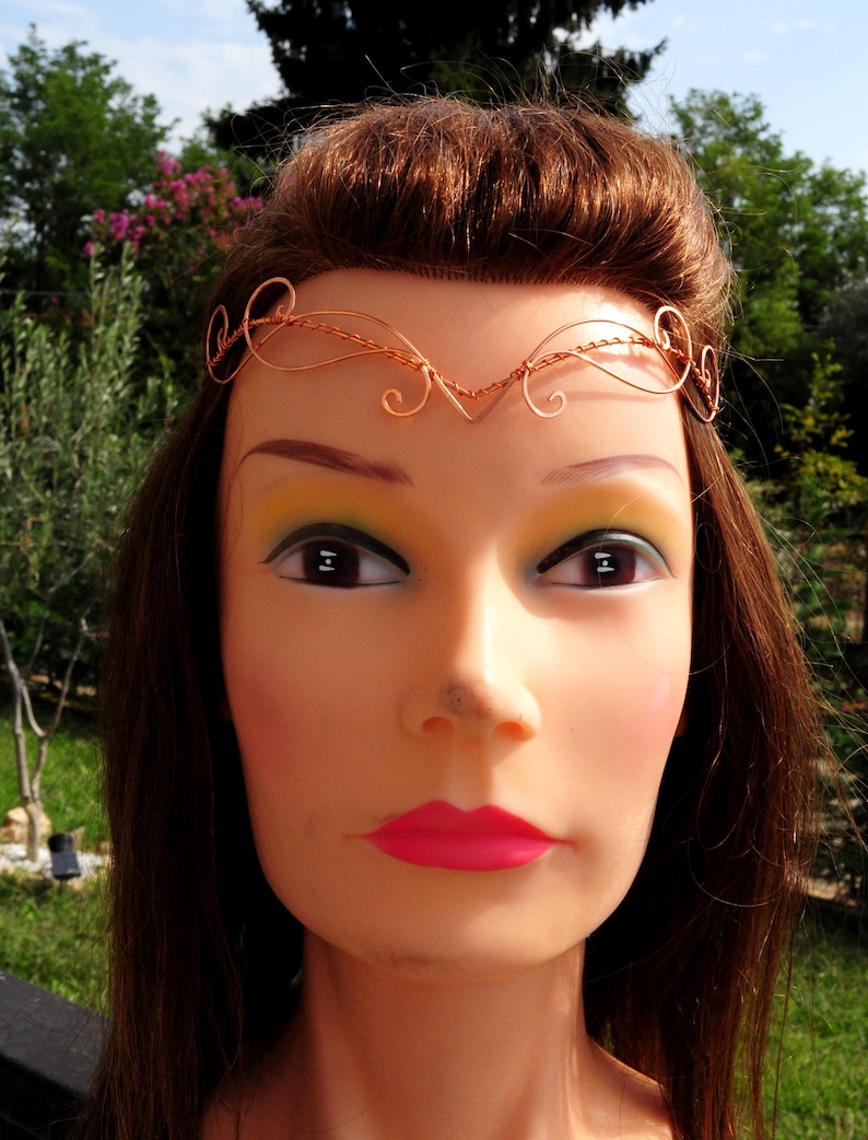 May include: A copper wire headband with a delicate, swirling design. The headband is worn on a mannequin with long brown hair and pink lipstick.