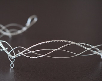 May include: A silver wire crown with a delicate, intricate design. The crown is made of multiple strands of wire that are twisted and intertwined to create a flowing, organic shape.