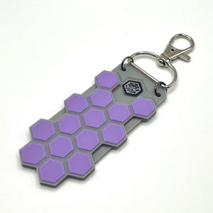 May include: A gray and purple hexagonal keychain with a silver metal clasp. The keychain has a small black logo in the center.
