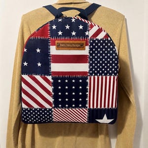May include: A backpack featuring a patchwork design of red, white, and blue stars and stripes. The fabric panels are joined with visible white stitching. A rectangular brown leather label with the text Daisy Muse Designs is centered on top.