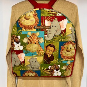 May include: A colorful backpack featuring a patchwork design with various jungle animals, including lions, giraffes, and elephants. The backpack has red straps and trim, and the text "Daisy Muse Designs" is visible on the front.