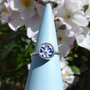 May include: A silver ring featuring a circular ceramic design in white and blue. The ring is presented on a light blue cone, with white and pink flowers in the background. The ring's design is reminiscent of Delftware.