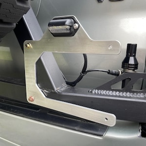 May include: A silver-colored metal bracket with an attached black LED light, mounted on the rear of a vehicle. The bracket is secured with bolts and has a sleek, angular design. A black cylindrical component is also visible.