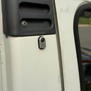 May include: A white vehicle exterior panel featuring a black plastic vent with three horizontal slots secured by a screw. Below the vent is a small, rectangular silver metal plate with a black rubber border, fastened by a single black bolt.
