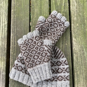 May include: A set of grey and brown knitted fingerless gloves and a matching hat with a geometric diamond pattern. The gloves and hat are made of a soft, textured yarn, displayed on a weathered wooden surface.