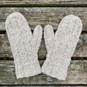 May include: A pair of light gray knitted mittens with a cable knit design. The mittens are made of a soft, textured yarn and are displayed on a weathered wooden surface. The mittens are designed to keep hands warm.