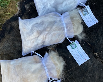 May include: Three bags of French Angora fiber, each filled with soft, fluffy wool in shades of white and beige. The bags are tied with white ribbons and have small tags with text. The fiber is displayed on a tree stump.