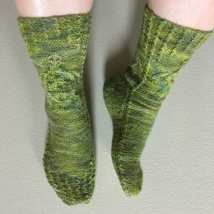 May include: A pair of hand-knitted socks in shades of green and yellow. The socks feature a textured pattern on the leg and foot, with a detailed design on the front of the leg. The yarn has a variegated color effect.