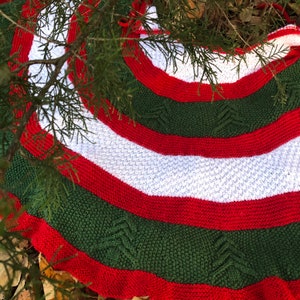 May include: A red, white, and green knitted Christmas tree skirt with a cable knit pattern. The skirt is decorated with a repeating pattern of stylized Christmas trees.
