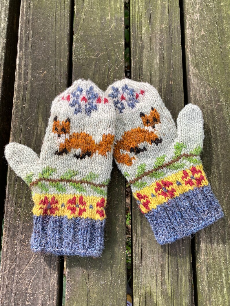 Puede incluir: Un par de guantes tejidos con fondo blanco y un patr&oacute;n colorido de zorros, flores y copos de nieve. Los guantes tienen un pu&ntilde;o azul.