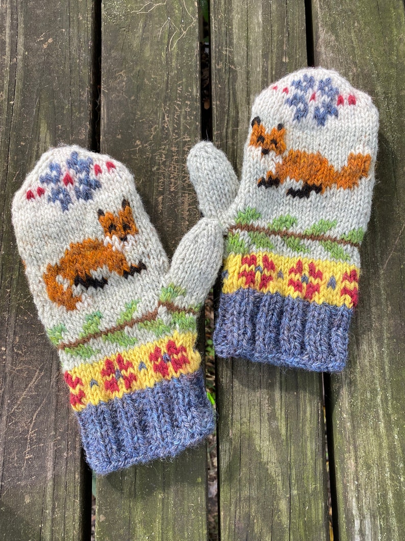 Puede incluir: Un par de guantes tejidos con un dise&ntilde;o de zorro. Los guantes son grises con detalles amarillos, rojos y verdes. Los pu&ntilde;os son azules.