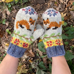 Puede incluir: Un par de guantes tejidos con un dise&ntilde;o de zorro. Los guantes son blancos con un patr&oacute;n verde, amarillo y rojo. Los pu&ntilde;os son azules.