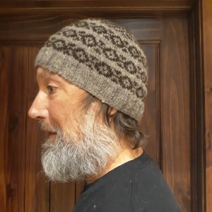 Puede incluir: Un gorro de punto gris con un patrón geométrico en marrón oscuro. El sombrero lo lleva una persona con barba larga y gris y una camisa oscura. El fondo es una puerta de madera.