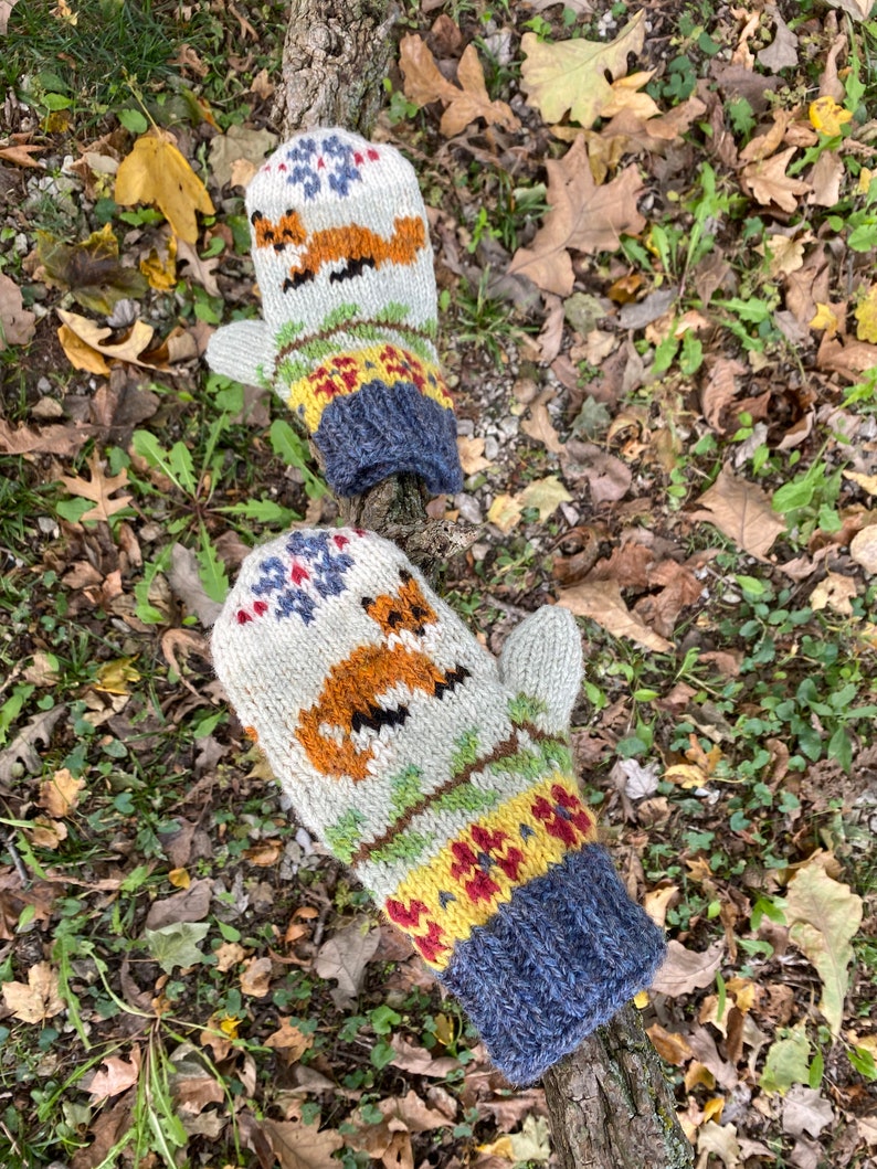 Puede incluir: Un par de guantes tejidos con un dise&ntilde;o de zorro. Los guantes son blancos con detalles en naranja, amarillo, rojo y azul. Los guantes est&aacute;n sobre una rama con hojas ca&iacute;das.