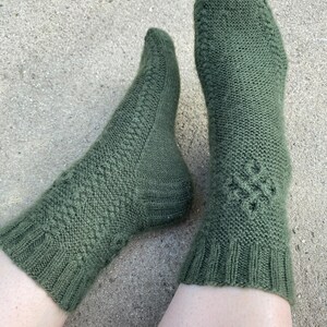 Op de afbeelding: Een paar olijfgroene gebreide sokken. De sokken hebben een geribbelde boord, een textuurpatroon op de voet en een decoratief ontwerp aan de bovenkant. Gemaakt van een zacht, warm materiaal.