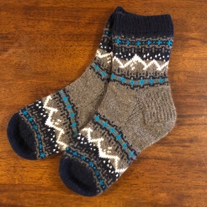 May include: A pair of grey knitted socks with a navy blue cuff, heel, and toe. The socks feature a geometric pattern in white and light blue. The socks are made of a soft, warm material, ideal for cold weather.