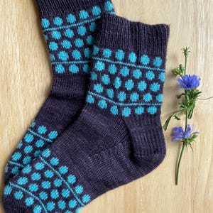 May include: A pair of dark purple knit socks with a repeating pattern of turquoise polka dots. The socks are displayed on a light wooden surface, with a sprig of blue flowers to the side.