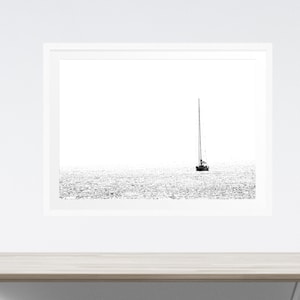 K&ouml;nnte beinhalten: Schwarz-Wei&szlig;-Druck eines Segelboots auf dem Wasser. Das Bild ist eine minimalistische Seelandschaft, mit dem Boot in der Mitte und dem Wasser, das das Licht reflektiert. Der Druck wird an einer wei&szlig;en Wand &uuml;ber einem hellen Holztisch angezeigt.