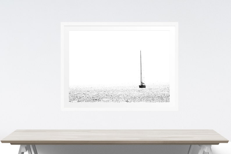 K&ouml;nnte beinhalten: Schwarz-Wei&szlig;-Druck eines Segelboots auf dem Wasser. Das Bild ist eine minimalistische Seelandschaft, mit dem Boot in der Mitte und dem Wasser, das das Licht reflektiert. Der Druck wird an einer wei&szlig;en Wand &uuml;ber einem hellen Holztisch angezeigt.