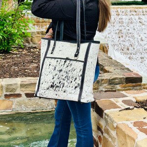 May include: A white and black cowhide tote bag with black leather trim and straps. The bag has a large main compartment and a smaller pocket on the front.