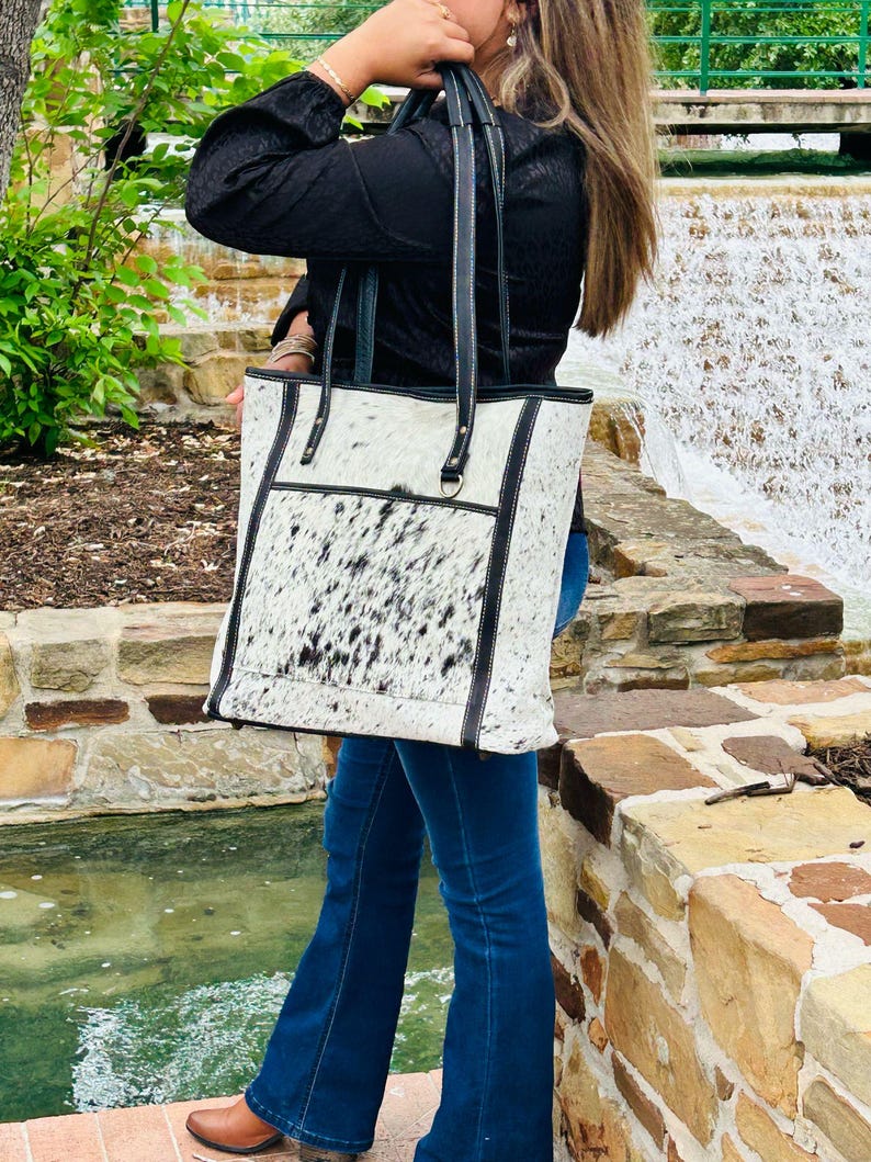 May include: A white and black cowhide tote bag with black leather trim and straps. The bag has a large main compartment and a smaller pocket on the front.