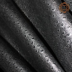 May include: Close-up of black leather with a textured, embossed pattern. The leather has a subtle sheen and a slightly raised, circular pattern.