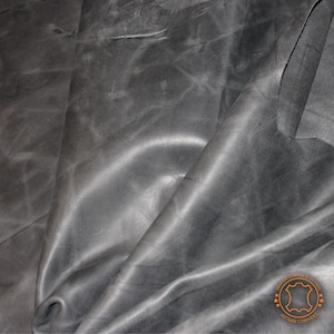 Puede incluir: Primer plano de una piel de vaca de cuero gris oscuro con textura arrugada. El cuero está doblado y drapeado, mostrando su flexibilidad. Un logotipo circular con el texto "PREMIUM COWHIDE & LEATHER ARTISAN COWHIDES" es visible en la esquina inferior derecha.