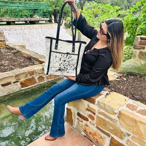 May include: A white and black cowhide tote bag with black leather trim and handles. The bag has a zippered pocket on the front.