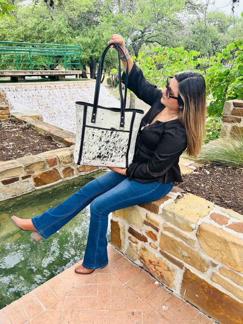 May include: A white and black cowhide tote bag with black leather trim and handles. The bag has a zippered pocket on the front.