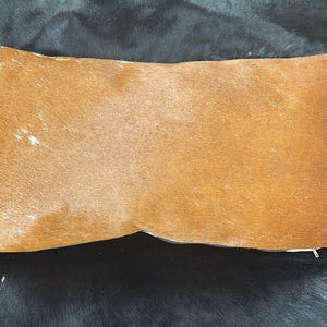 Pode incluir: Uma almofada decorativa retangular com uma superfície castanha texturizada. A almofada tem um fecho de correr e está sobre um fundo escuro e texturizado. A cor da almofada é um tom quente e terroso.