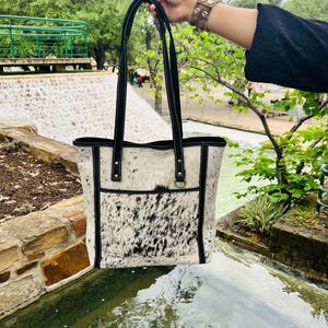 May include: A white and black cowhide tote bag with black leather straps and trim. The bag has a large pocket on the front and a small pocket on the inside.
