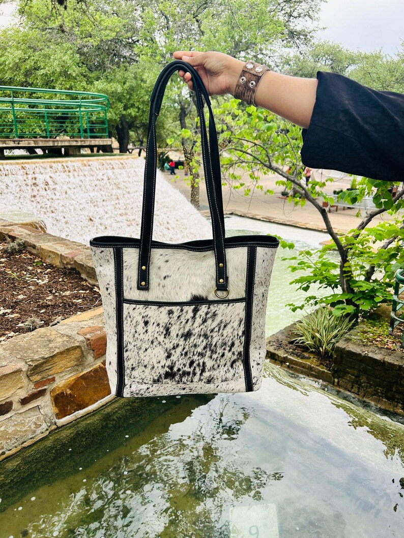 May include: A white and black cowhide tote bag with black leather straps and trim. The bag has a large pocket on the front and a small pocket on the inside.