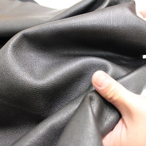 K&ouml;nnte beinhalten: Nahaufnahme von schwarzem Leder mit einer genarbten Textur. Das Leder ist weich und geschmeidig und wird von einer Hand gehalten.
