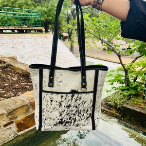 May include: A black and white cowhide tote bag with black leather trim and straps. The bag has a front pocket and a gold metal ring.