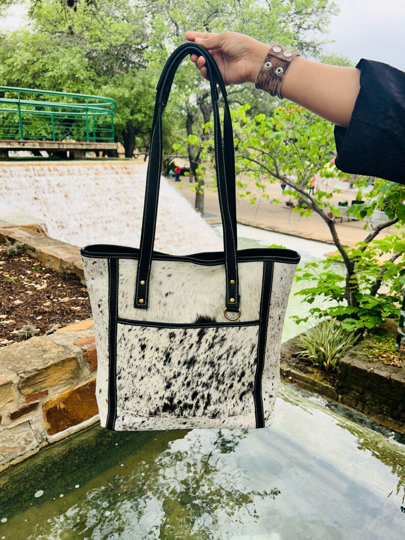May include: A black and white cowhide tote bag with black leather trim and straps. The bag has a front pocket and a gold metal ring.