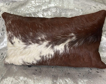 May include: Rectangular cowhide pillow with a brown and white pattern. The pillow has a natural, textured appearance, with the brown sections dominating the design. The white section is in the center of the pillow.